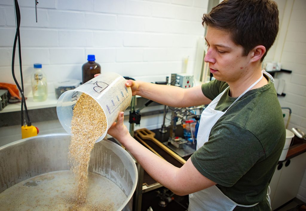 Für den ersten Schritt des Brauprozessen, das Maischen, wird Malz in Wasser gekocht. Niklas Hoffmann schüttet den ersten Messbecher hinein. Foto: Universität Bielefeld / Mike-Dennis Müller