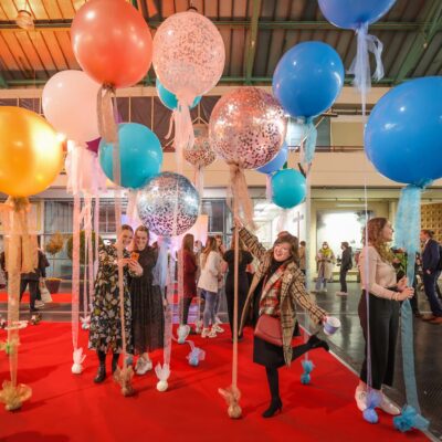 Tag für Absolvent:innen: Die besondere bute Luftballonkulisse auf rotem Teppich in der Uni-Halle loggt zahlreiche Absolvent:innen zum Fotos machen an.